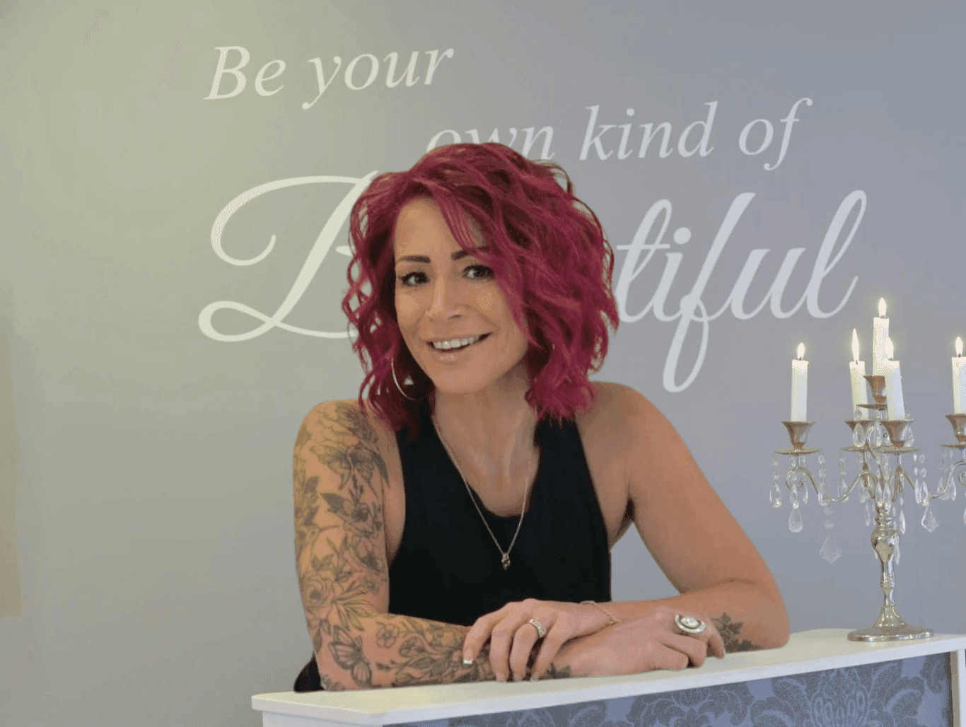 Annabelle Fontaine, owner of The Dolls House Hairdressing and Beauty Boutique in Eastbourne, smiling at the salon reception desk with pink hair, floral tattoos, and a wall sign reading “Be your own kind of beautiful” in the background.
