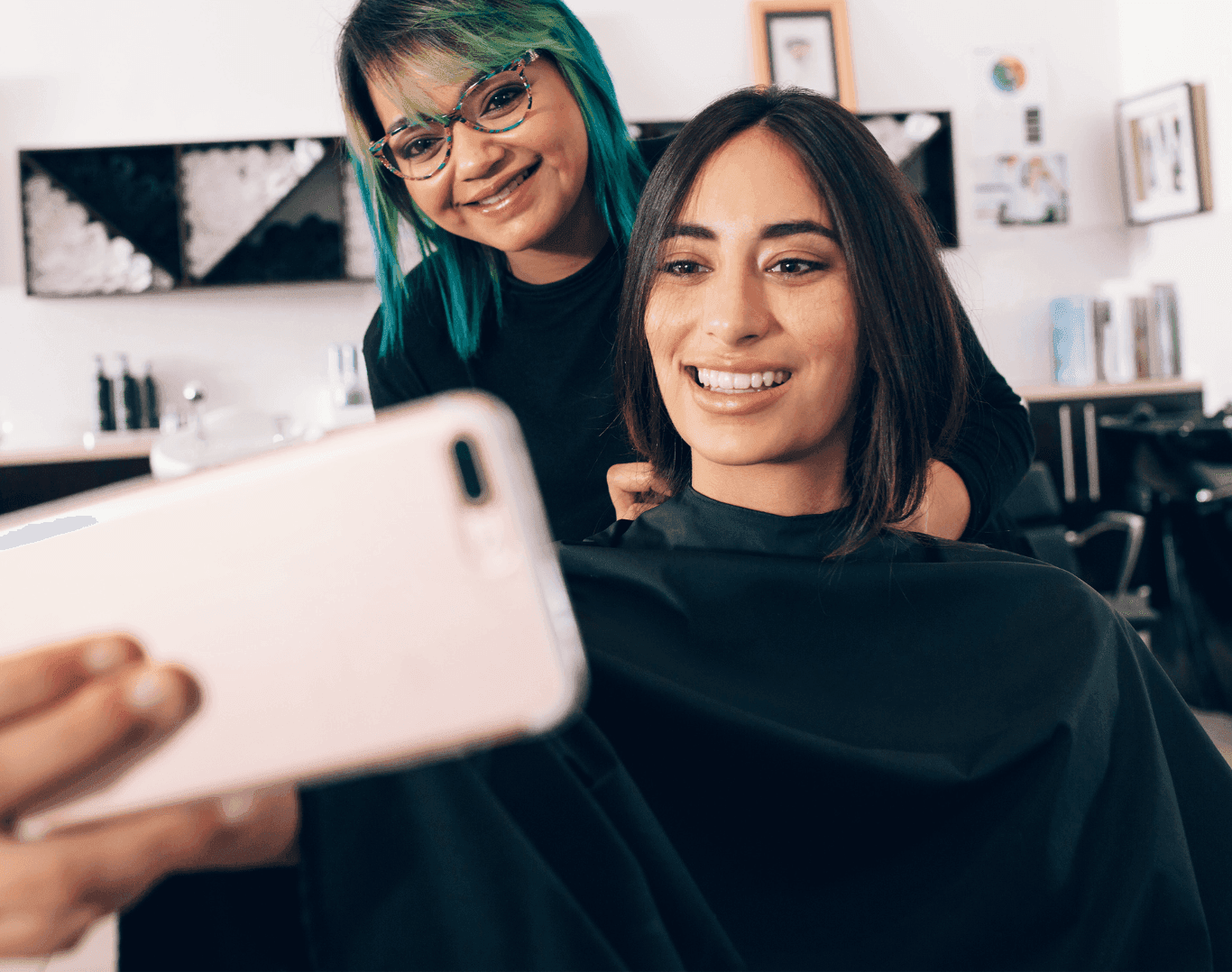 Smiling hairstylist with colourful hair helping a client take a selfie in the salon after a haircut.