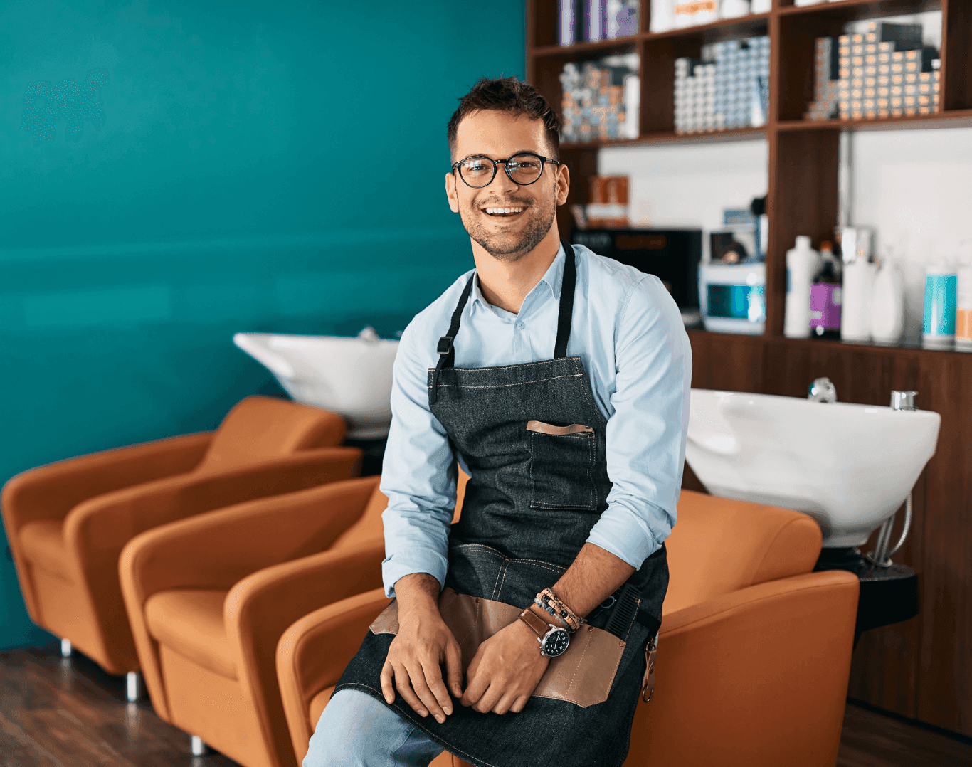 Smiling salon owner wearing an apron, sitting confidently in front of salon backwash chairs, ready to welcome clients.