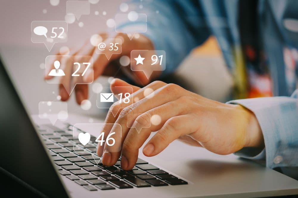 Hands typing on a laptop with floating social media icons and metrics like likes, comments, messages, and followers, representing performance review.