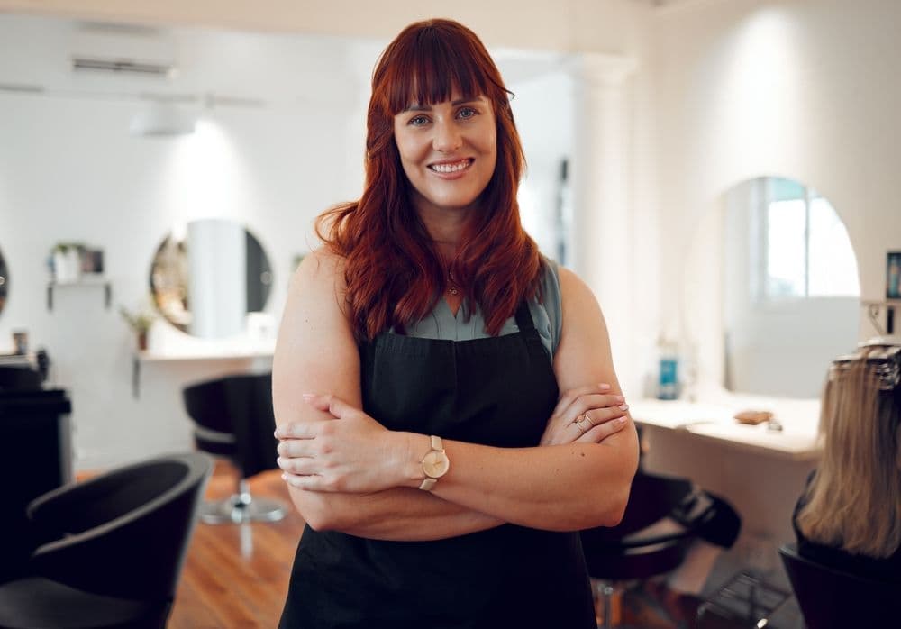 Confident hairstylist standing with folded arms inside a modern salon, symbolising setting clear benchmarks for social media success.