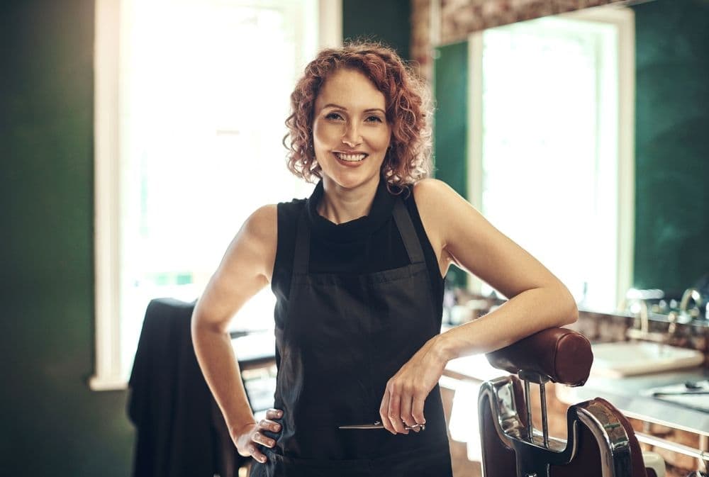 Confident salon owner smiling inside a modern salon with floating social media icons and booking confirmations, representing posts that turn engagement into real appointments.