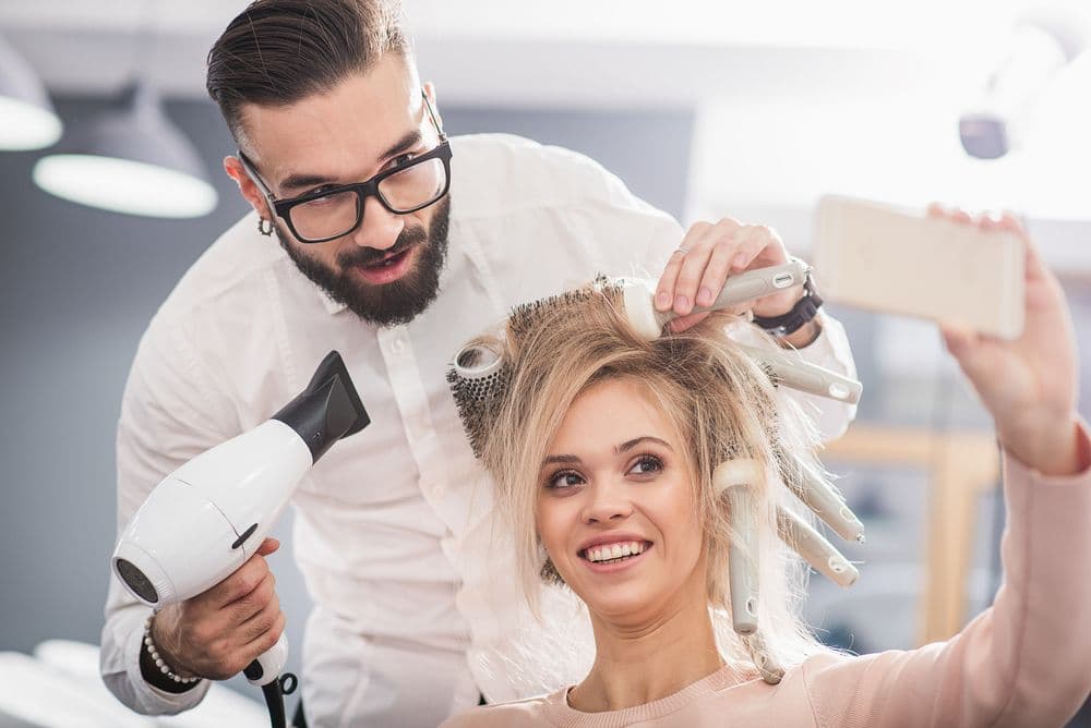 Smiling salon client taking a selfie while a stylist blow dries and styles her hair, capturing authentic client-generated content.