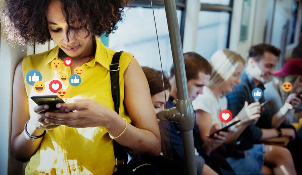 Young woman smiling while using her phone with floating social media reaction icons, surrounded by others also engaging on their devices, representing online community building.