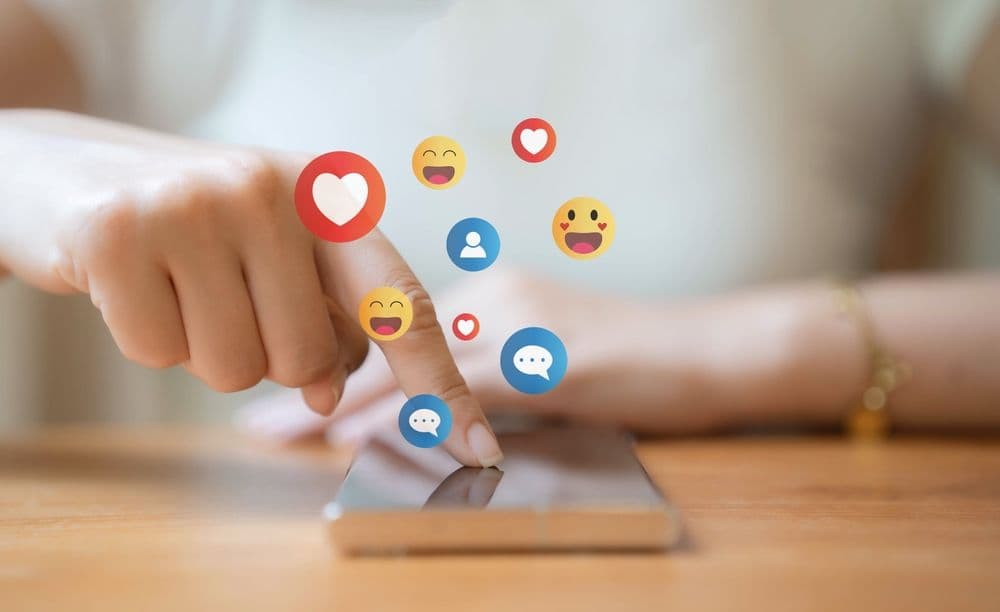 Close-up of a person using a smartphone with floating social media icons like hearts, comments, and smiley faces representing engagement.