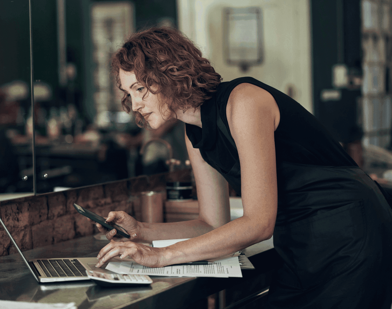 Hair salon owner looking at laptop and smartphone, planning her website strategy and digital marketing to improve online bookings and Google search rankings.